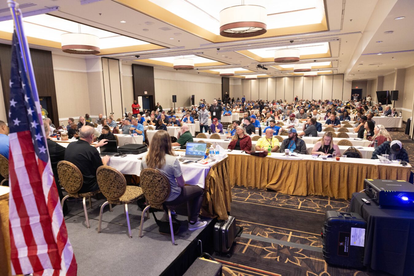 A group of delegates in a large conference room at tables with gold table cloths. In the foreground is an American flag